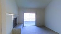 This single-floor space centers around a comfortable living area defined by soft carpeted flooring and neutral-toned walls that create a clean, understated aesthetic. A standout feature is the tile-surround fireplace positioned along one wall, framed in light-colored tile with a classic raised hearth, adding both visual interest and a functional focal point to the room. Opposite the fireplace, a sliding glass door fitted with vertical blinds opens to an outdoor balcony, allowing natural light to fill the interior and providing access to an exterior view. The vaulted ceiling height contributes to an open, airy atmosphere, enhancing the sense of spaciousness within the living area. The overall design is straightforward and practical, with minimal ornamentation, offering a neutral canvas suitable for a variety of furnishing styles. The combination of the fireplace, natural light from the sliding door, and the elevated ceiling makes the living space feel inviting and well-proportioned.