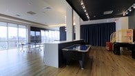 This single-floor recreation space offers a thoughtfully designed entertainment environment that blends modern aesthetics with a dynamic, social atmosphere. The open-plan layout is anchored by warm hardwood flooring throughout, which contrasts beautifully with the bold black ceiling accents and crisp white walls. A striking half-wall partition divides the space into distinct activity zones while maintaining an airy, connected feel. An air hockey table takes center stage in the primary zone, complemented by a ping pong table positioned near the expansive floor-to-ceiling windows. These large windows flood the space with natural light and frame impressive elevated city views, enhancing the sense of openness. Dramatic track lighting with directional spotlights runs along the dark ceiling, providing precise illumination across the activity areas. Globe-style vanity lights add a theatrical touch to the partition wall, contributing to the venue's polished character. Deep navy blue curtains line the walls, offering a rich, sophisticated backdrop that can be drawn for privacy or atmosphere control. The overall ambiance is sleek and modern, making this space well-suited for recreational use, social gatherings, or team-building activities in an upscale urban setting.