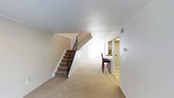 This two-story space is designed with a straightforward, functional layout that maximizes usability across both levels. The main floor features neutral, light-toned carpeting that runs throughout the open area, creating a cohesive and understated aesthetic. A carpeted staircase with a classic wrought iron railing serves as a central architectural element, providing an elegant transition between floors while also anchoring the open layout visually.

The main living area is spacious and open, with crisp white walls and ceilings that reflect light and contribute to a clean, airy atmosphere. Toward the rear of the space, a built-in counter with bar-height seating offers a practical and versatile area, suitable for casual dining or use as a workspace. The counter opens into an adjacent room, hinting at a connected kitchen or utility space beyond.

Natural light filters in through what appears to be a window at the far end of the space, brightening the interior and enhancing the sense of openness. The overall design is neutral and well-maintained, with consistent trim and baseboards throughout. The space beneath the staircase is neatly enclosed, making efficient use of the available square footage. The two-floor layout offers both communal and private areas, making the space adaptable to a variety of uses.