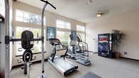 This single-level fitness room offers a well-organized workout environment designed to accommodate a range of exercise needs. The space is floored with neutral carpet throughout, transitioning to interlocking rubber mat tiles in the designated strength and free-movement area, providing appropriate surface support for varied activities. Along one wall, a large floor-to-ceiling mirror expands the sense of space and allows users to monitor their form during exercise.

Cardio equipment is the focal point of the room, featuring multiple treadmills—including a Star Trac model—alongside several elliptical cross-trainers arranged to face a bank of large, multi-pane windows. These windows admit generous natural light and offer views of surrounding trees and the outdoor environment, creating an open and pleasant workout atmosphere. Transom-style windows positioned above the main window line further enhance brightness throughout the room.

A pull-up and dip tower station sits on the rubber mat zone, supporting bodyweight strength training. Additional amenities include a standing scale, framed instructional fitness posters, a decorative potted plant, and a Pepsi beverage vending machine positioned near the entry door. The textured ceiling, ceiling fan, and flush-mount lighting complete the functional and straightforward interior of this fitness facility.