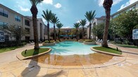 This single-story amenity space is designed to evoke a resort-like atmosphere, anchored by a generously sized freeform swimming pool with a shallow wading entry and striking turquoise water. Tall palm trees frame the pool on all sides, rising above raised landscaped planters that add lush greenery to the hardscaped surroundings. The pool deck is finished in warm-toned flagstone, extending outward to a spacious patio area furnished with lounge chairs and shaded by blue fabric umbrellas.

The adjacent clubhouse is constructed of natural limestone masonry, with arched openings and large floor-to-ceiling glass walls that visually connect the interior to the outdoor space. Inside, soaring vaulted ceilings with exposed structural elements and an oversized ceiling fan create an airy, open environment. Wicker lounge furniture with navy and white accent pillows is arranged throughout the interior, encouraging relaxed social gatherings.

A substantial outdoor limestone fireplace is integrated into the covered corridor, providing a focal gathering point during cooler months. The covered porch area transitions seamlessly between indoors and outdoors through frameless sliding glass panels. Throughout, the consistent use of flagstone flooring, natural stone walls, and curated landscaping creates a cohesive, upscale ambiance.