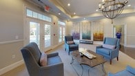 This single-floor community lounge space is thoughtfully designed to offer both comfort and elegance. The layout flows seamlessly across two connected seating areas, each characterized by warm neutral tones, plush carpeting, and rich hardwood flooring that anchors the transition between zones. Tray ceilings with recessed lighting and crown molding add architectural distinction, while a chandelier provides a refined finishing touch. The main lounge area features a curated arrangement of upholstered armchairs and a wooden coffee table, creating an inviting space for conversation and relaxation. A large-scale, multi-panel abstract artwork in soft blues, purples, and warm earth tones serves as a striking focal point along one wall. The adjacent area offers additional seating alongside a wall-mounted flat-screen television and a console table adorned with decorative vases and greenery. Throughout the space, oversized French doors with transom windows line the exterior walls, flooding the interior with natural light and offering views of the surrounding outdoor grounds. Decorative round mirrors mounted above dark-finished sideboards reflect light and add depth to the entryway corridor, which connects the two primary gathering areas with an open, airy feel. The overall ambiance is polished yet welcoming, making this a versatile space suited for both quiet retreat and community engagement.