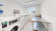 Located at 3388 Bathurst Street in Toronto, this shared laundry facility offers a clean, well-maintained space designed for efficient, practical use. The room features a row of commercial-grade front-loading washing machines paired with top-loading dryers, providing ample capacity for residents. A large stainless steel work table runs along one side of the room, offering generous space for sorting and folding laundry. A utility sink is also available, positioned conveniently near the machines. The walls are finished with white shiplap-style tile wainscoting topped by smooth white-painted drywall, creating a bright and tidy atmosphere. The floor is laid in a distinctive light-toned wood-look tile in a herringbone pattern, adding a refined touch to the functional space. Fluorescent strip lighting illuminates the room evenly, while a window along one wall allows in natural light. Safety is addressed with a visible fire extinguisher mounted near the entrance. An information board is posted on the wall, providing usage guidelines for residents. Access is through a metal-framed door that connects to the building's common corridor. The overall design is straightforward and functional, prioritizing cleanliness and ease of use.