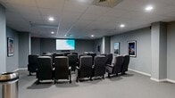 Located at 1099 N McMullen Booth Rd in Clearwater, Florida, this dedicated private screening room delivers an authentic cinema experience within a compact, well-appointed single-floor layout. The space is designed exclusively for movie viewing, with rows of dark upholstered theater-style seats — each equipped with armrests and cup holders — arranged to provide clear sightlines to the large wall-mounted display screen at the front of the room. A soundbar is mounted beneath the screen, supporting an immersive audio experience. The walls are painted in a neutral gray tone, creating a focused, cinema-like atmosphere, and are adorned with framed classic movie posters, including recognizable titles that reinforce the theatrical theme. Recessed ceiling lighting and a suspended tile ceiling complete the polished interior. A built-in concession alcove along one wall houses a popcorn machine and counter space, adding a fun, functional touch. Carpeted flooring throughout contributes to both comfort and acoustic performance. Structural columns are integrated into the layout without impeding the viewing area. A clearly marked exit door ensures safety compliance. This thoughtfully designed screening room is well-suited for private film screenings, events, or entertainment programming.