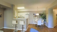 This single-floor residence showcases an open-concept layout that seamlessly connects the kitchen, breakfast bar, and dining area into one cohesive living space. The kitchen is outfitted with white cabinetry, white appliances including a built-in microwave and refrigerator, and light countertops that contribute to a clean, bright aesthetic. A peninsula-style breakfast bar extends from the kitchen, providing additional seating with two modern white bar stools featuring chrome bases — ideal for casual dining or entertaining.

Adjacent to the kitchen, the dining area is anchored by a round table surrounded by sleek white chairs with chrome legs. Above the table hangs a contemporary pendant light fixture that adds a refined focal point to the space. A decorative mosaic-style mirror on the wall and a large indoor plant add visual interest and a touch of warmth to the area.

The warm hardwood-style flooring runs throughout, contrasting pleasantly with the soft neutral gray and white wall tones. The overall ambiance is modern and airy, with a cohesive color palette of whites, grays, and warm wood tones that creates an inviting and functional living environment.