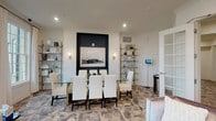 Located at 200 W Main St in New Albany, Ohio, this thoughtfully designed single-floor space exudes a polished, contemporary aesthetic. The interior is anchored by a striking dining area showcasing a glass-top table surrounded by crisp white upholstered chairs, creating an inviting yet refined atmosphere. A bold dark accent wall serves as a dramatic focal point, framing a large landscape artwork flanked by elegant wall sconces that cast a warm, ambient glow. The flooring features an intricate geometric parquet-style pattern in mixed wood tones, adding visual depth and a sense of artisanal craftsmanship underfoot. Flanking the dining area are tall, open brass-framed shelving units displaying curated décor pieces, books, and accessories that contribute to the space's cultivated character. The crisp white walls and recessed ceiling lighting create a bright, clean backdrop that complements the sophisticated furnishings. A glimpse into the adjacent area reveals a frosted glass-paneled door, suggesting additional functional spaces beyond. The overall ambiance is one of modern elegance — refined, well-appointed, and meticulously curated, making this New Albany location a distinguished and memorable space.