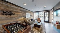 Located at 1005 W Main St in Durham, NC, this single-floor amenity space blends industrial-modern design with warm, inviting elements throughout. The lounge area is anchored by a striking reclaimed wood accent wall that spans the length of the room, providing rich texture and a rustic contrast to the clean, contemporary finishes. Comfortable seating — including a neutral sofa, leather accent chairs, and a bold geometric area rug — creates an inviting gathering space, while a foosball table adds a playful, social element. Large floor-to-ceiling glass doors and sliding panels flood the interior with natural light and open directly to an outdoor courtyard area, seamlessly connecting the indoor and outdoor spaces. An adjoining dining and entertaining area features a substantial solid wood communal table with industrial-style seating, pendant lighting, and a stone tile accent wall framing a wet bar with cabinetry and under-counter appliances. A large glass overhead door in this section further enhances the indoor-outdoor flow. Recessed lighting, large-format tile flooring, and curated artwork displayed throughout contribute to the cohesive, upscale aesthetic. The space is well-suited for both casual socializing and private events.