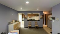 This single-floor space offers a functional and straightforward layout designed for practicality and ease of movement. The main living area features wall-to-wall carpeting in a neutral tone and is painted in a calming blue-gray hue that carries throughout the space, lending a cohesive and understated aesthetic.

A defining feature of this space is the pass-through kitchen window, which connects the kitchen to the main living area and promotes an open, communicative flow between rooms. The kitchen is equipped with warm wood cabinetry, a white refrigerator, a microwave, and a dark countertop, with recessed lighting overhead providing consistent illumination. A bar-height counter with seating for two chairs invites casual dining or conversation directly from the living area.

Adjacent to the kitchen, a separate utility or laundry area is visible through a doorway, housing white appliances and additional counter space. Recessed ceiling lights throughout the main area ensure even, practical lighting. To one side, a hallway leads to additional rooms, suggesting further private or functional spaces beyond what is immediately visible.

Decorative touches include floral artwork on the walls, adding warmth to the otherwise utilitarian setting. A storage unit with filing drawers anchors one corner of the main room, hinting at a multi-purpose use for this versatile space.