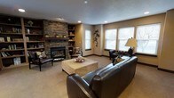 This single-floor space centers around a beautifully appointed living room that immediately draws attention with its floor-to-ceiling stacked-stone fireplace, serving as the room's defining focal point. The natural texture of the stonework adds depth and character, flanked on either side by warm wood built-in bookshelves that provide both functional storage and display space for decorative accents and books. Recessed lighting in the ceiling complements the natural light that filters in through multiple windows dressed with clean white horizontal blinds, creating a bright yet cozy ambiance throughout. The neutral, earthy tones of the walls pair seamlessly with the plush wall-to-wall carpeting, lending the space a grounded, comfortable feel. The seating area is thoughtfully arranged around a woven square coffee table, anchored by rich dark leather sofas and accent chairs that add a touch of sophistication. Decorative wall art and a classic table lamp contribute to the room's layered, well-curated aesthetic. Overall, the space strikes a balance between refined design and everyday comfort, making it an ideal setting for relaxing or entertaining.