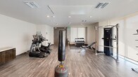 This fitness center is a thoughtfully designed, single-floor space that offers a comprehensive range of exercise equipment in a clean, modern setting. The room features wood-look tile flooring throughout, with rubberized matting in designated workout zones to provide additional cushioning and traction. Bright white walls and recessed lighting create a well-lit, open atmosphere.

The cardio area is anchored by multiple True-brand treadmills and elliptical machines, complemented by a large flat-screen TV mounted above a dark wood media console — ideal for entertainment during workouts. A freestanding punching bag occupies a central position in the room, adding a versatile option for conditioning and combat training.

The strength training section includes a multi-functional cable machine, an adjustable weight bench, and a well-stocked dumbbell rack featuring a range of weights neatly organized on a tiered stand. A stretch and mat area with rubber flooring provides additional space for flexibility training or floor exercises. A bold framed art piece adds a decorative focal point to the space.

The facility is accessed through wide double doors and includes a window with roller shades that allows for natural light while maintaining privacy. Ceiling-mounted HVAC vents ensure comfortable airflow throughout, making this fitness center a functional and inviting environment for a full-body workout.