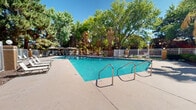 Located at 9300 Montana Ave in El Paso, Texas, this outdoor pool area offers a well-maintained and inviting recreational environment suited for warm desert days. The centerpiece of the space is a generously sized swimming pool with clearly marked depth indicators, ranging from a shallow entry point to a deeper section, ensuring safety for a variety of swimmers. The pool features sturdy stainless steel entry railings for convenient access and is finished with vivid turquoise water that reflects the ample Texas sunlight.

The surrounding pool deck is constructed of smooth concrete, providing ample space for lounge chairs and seating arrangements positioned to take full advantage of the sun and shade. A white metal security fence encloses the entire pool area, adding both safety and a clean, defined perimeter to the space.

What distinguishes this outdoor area is the remarkable natural canopy formed by a dense collection of mature trees, including towering palms and broad-leafed shade trees, which lend a lush, verdant atmosphere uncommon in the El Paso desert landscape. Shaded seating areas with umbrellas and chairs are tucked along the perimeter, offering comfortable spots to relax away from direct sunlight.

An adjacent single-story building with a terracotta-toned exterior and tiled roof serves as a functional backdrop, likely housing support amenities for the pool area. A decorative circular hardscape feature with surrounding seating adds additional character to the outdoor space.
