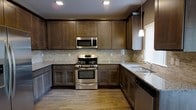 This two-story home showcases a thoughtfully designed kitchen as one of its standout features. The kitchen is outfitted with rich, dark-stained shaker-style cabinetry that extends to the ceiling, providing generous storage space. Granite countertops in a light, speckled finish complement the cabinetry, offering both durability and visual contrast. Stainless steel appliances, including a gas range, over-the-range microwave, and dishwasher, lend a modern, cohesive aesthetic to the space. A textured stone tile backsplash adds depth and dimension to the walls, tying together the darker tones of the cabinetry with the lighter countertops. The kitchen also features a stainless steel sink positioned near a window, allowing for natural light to fill the workspace. Warm hardwood-style flooring runs throughout, enhancing the overall inviting atmosphere. Recessed lighting and a pendant light above the sink area provide layered illumination. The layout follows a functional U-shape design, maximizing counter and storage space. Spanning two floors, this home offers versatile living arrangements suited to a variety of lifestyles.