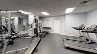 This fitness center offers a fully equipped workout environment designed to support a wide range of exercise routines. The space is laid out across a single floor with a practical, open floor plan that allows for comfortable movement between stations and equipment.

The room features light grey walls, a drop ceiling with recessed panel lighting, and durable dark carpet flooring suited for a high-traffic fitness environment. Large wall-to-wall mirrors line one side of the space, visually expanding the room and allowing users to monitor their form during workouts.

On the cardio side, the facility includes multiple treadmills from Life Fitness, as well as elliptical cross-trainers, providing solid options for aerobic exercise. A rowing machine is also available for full-body cardiovascular conditioning.

The strength training area is anchored by a Schwinn multi-station cable machine, accommodating a variety of resistance exercises. Complementing this is a dedicated free weights section featuring a tiered dumbbell rack stocked with a range of weights, along with flat workout benches for pressing and accessory movements.

The space is well-lit through a combination of recessed and flush-mounted ceiling lights, creating a bright and energizing atmosphere. The overall design is clean and utilitarian, prioritizing function and accessibility to deliver a complete fitness facility experience.