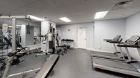 This fitness center offers a fully equipped workout environment designed to support a wide range of exercise routines. The space is laid out across a single floor with a practical, open floor plan that allows for comfortable movement between stations and equipment.

The room features light grey walls, a drop ceiling with recessed panel lighting, and durable dark carpet flooring suited for a high-traffic fitness environment. Large wall-to-wall mirrors line one side of the space, visually expanding the room and allowing users to monitor their form during workouts.

On the cardio side, the facility includes multiple treadmills from Life Fitness, as well as elliptical cross-trainers, providing solid options for aerobic exercise. A rowing machine is also available for full-body cardiovascular conditioning.

The strength training area is anchored by a Schwinn multi-station cable machine, accommodating a variety of resistance exercises. Complementing this is a dedicated free weights section featuring a tiered dumbbell rack stocked with a range of weights, along with flat workout benches for pressing and accessory movements.

The space is well-lit through a combination of recessed and flush-mounted ceiling lights, creating a bright and energizing atmosphere. The overall design is clean and utilitarian, prioritizing function and accessibility to deliver a complete fitness facility experience.