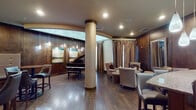 Located at 5290 Big Island Dr in Jacksonville, FL, this sophisticated single-floor lounge and bar space exudes warmth and elegance through its carefully curated design. Rich dark wood paneling lines the walls throughout, creating a cohesive and upscale atmosphere that defines the entire space.

A grand piano anchors one section of the lounge, positioned beneath a striking arched ceiling detail with soft backlighting that adds an artistic focal point. Textured decorative columns rise from the hardwood floors, complementing the warm tones of the interior. Contemporary landscape artwork adorns the walls, adding refined visual interest.

The seating area offers plush wingback chairs and cushioned armchairs arranged for intimate conversation, accompanied by ottomans and side tables. Tall draping curtains frame the windows, softening the natural light that filters into the space.

A dedicated dining and bar lounge area features high-top bistro tables paired with leather-upholstered bar stools and tufted booth banquette seating along the walls. Elegant drum pendant lights illuminate this section, enhancing its club-like atmosphere.

A curved bar counter with granite surfaces and teardrop pendant lights above serves as the social hub of the space. Bar-height seating lines the counter alongside additional lounge chairs, creating an inviting setting for guests to gather and unwind.
