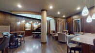 Located at 5290 Big Island Dr in Jacksonville, FL, this sophisticated single-floor lounge and bar space exudes warmth and elegance through its carefully curated design. Rich dark wood paneling lines the walls throughout, creating a cohesive and upscale atmosphere that defines the entire space.

A grand piano anchors one section of the lounge, positioned beneath a striking arched ceiling detail with soft backlighting that adds an artistic focal point. Textured decorative columns rise from the hardwood floors, complementing the warm tones of the interior. Contemporary landscape artwork adorns the walls, adding refined visual interest.

The seating area offers plush wingback chairs and cushioned armchairs arranged for intimate conversation, accompanied by ottomans and side tables. Tall draping curtains frame the windows, softening the natural light that filters into the space.

A dedicated dining and bar lounge area features high-top bistro tables paired with leather-upholstered bar stools and tufted booth banquette seating along the walls. Elegant drum pendant lights illuminate this section, enhancing its club-like atmosphere.

A curved bar counter with granite surfaces and teardrop pendant lights above serves as the social hub of the space. Bar-height seating lines the counter alongside additional lounge chairs, creating an inviting setting for guests to gather and unwind.