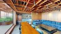 This thoughtfully designed urban amenity space seamlessly blends an indoor lounge with an expansive rooftop terrace, offering residents a sophisticated retreat above the city. The interior lounge is immediately distinguished by its dramatic wood lattice ceiling — a grid of warm, richly toned timber that creates an architectural focal point overhead. Wood-paneled walls reinforce the warm aesthetic, while track lighting illuminates the space throughout. A long communal dining table with ample seating anchors the room, complemented by a vibrant blue sectional sofa and a sleek marble-topped coffee table. A kitchenette with subway tile backsplash, open shelving with greenery, and modern cabinetry provides functional support for gatherings. The adjoining rooftop terrace extends the living experience outdoors, featuring light composite decking and a continuation of the wood pergola structure that bridges indoors and out. Curved Corten steel planters brimming with lush greenery add organic texture throughout the terrace. White lounge chairs, dining sets, and wire accent chairs are arranged across the terrace to accommodate both relaxation and al fresco dining. Panoramic views of the surrounding urban skyline — including prominent high-rise towers — frame the space, making it an exceptional setting for both everyday enjoyment and social gatherings.