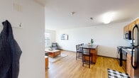 Located at 117 Washington Ave in Schenectady, NY, this well-appointed single-floor unit offers a clean, modern design throughout. The open-concept living and dining area is anchored by warm hardwood-style flooring and crisp white walls, creating a bright and welcoming atmosphere. Large windows flood the living room with natural light and frame views of the surrounding Schenectady cityscape, including vibrant street murals visible from multiple rooms. A wall-mounted TV and comfortable seating make the living area an ideal gathering space. Adjacent to the living room, a raised bar-height island with seating serves as a casual dining area. The kitchen features warm wood cabinetry, dark countertops, and black appliances, including a full-size refrigerator and range. The unit includes multiple bedrooms, each carpeted and equipped with closet storage. One room functions well as a dedicated home office, with a window overlooking the city. The bathroom offers a spacious dual-sink vanity with a large mirror, complemented by a separate tiled shower and toilet area. Throughout the unit, the consistent use of neutral tones, natural light, and functional layouts contribute to a cohesive and comfortable living environment.