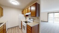 This single-floor, 2-bedroom, 1-bathroom space offers a well-organized and functional layout designed for efficient everyday living. The galley-style kitchen is a standout feature, lined on both sides with warm oak cabinetry offering generous storage, light-toned countertops, a double-basin sink, and white appliances including an electric range and refrigerator. Light wood-look flooring runs through the kitchen and hallway, adding a clean, cohesive aesthetic throughout the common areas. The central hallway connects all rooms seamlessly, with multiple doors providing access to the bedrooms, bathroom, and storage closets. The primary bedroom is notably spacious, featuring plush carpet, two closets — including a full bi-fold — and ample room for furnishings. The second bedroom is more compact but equally well-appointed, with carpet flooring, a ceiling light fixture, and a window that invites natural light. The bathroom is clean and straightforward, equipped with a full-size soaking tub and shower combo featuring a white surround with built-in shelving and chrome fixtures. Throughout the space, neutral wall tones, textured ceilings, and dark wood door and baseboard trim provide a consistent, understated design language.
