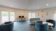 Located at 23641 Newhall Ave in Santa Clarita, CA, this single-story community lounge space is thoughtfully designed to serve as a functional and inviting gathering area. The room features a gently curved layout with smooth white walls and recessed lighting complemented by sleek pendant lights that add a modern touch to the interior.

A dedicated computer workstation area along one wall includes a long wooden desk equipped with dual monitors and stylish ghost-style chairs, making it a practical resource for residents. Adjacent to this, large windows and a set of French doors flood the space with natural light and offer views of the surrounding greenery, creating an open and airy atmosphere.

The seating areas are furnished with teal and blue upholstered chairs and curved sofas arranged to encourage conversation and relaxation. A small round white table anchors one of the lounge groupings, adding a clean, contemporary accent. Decorative ceramic wall art in warm orange and neutral tones provides a pop of color and artistic character throughout the space.

Soft carpet in a blue-grey hue covers the floors, contributing to the room's comfortable and cohesive aesthetic. A wall-mounted television and additional seating areas with French door access to an outdoor area further enhance the versatility of this well-appointed community space.