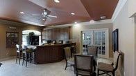 This single-floor space offers a well-appointed interior designed for both functionality and comfort. The main area serves as a central hub, featuring a large, curved service or reception counter finished in rich dark wood cabinetry that extends to the ceiling, providing ample storage and a polished, professional aesthetic. Recessed lighting and a ceiling fan keep the space bright and comfortable, while a tray ceiling with a warm accent color adds an architectural focal point overhead. Light-toned tile flooring runs throughout, contributing to a clean and open feel. A dining or meeting area sits adjacent to the counter, furnished with a glass-topped table and upholstered chairs, making it well-suited for gatherings or casual meetings. Classic white French doors lead to a secondary room, allowing natural light to filter through and offering a sense of connectivity between spaces. A window with an arched detail near the counter adds character and brings in additional daylight. The overall ambiance is warm and inviting, blending traditional design elements with practical amenities to create a versatile and welcoming environment.