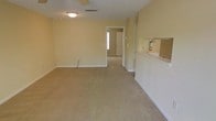 This single-story space features an open, rectangular layout finished in warm neutral tones throughout. The main living area is covered in wall-to-wall beige carpeting, lending a soft, understated feel to the room. Walls are painted in a light cream or pale yellow tone, contributing to a bright, airy atmosphere. Overhead, a ceiling-mounted light fixture provides central illumination, and a standard HVAC vent is integrated into the ceiling for climate control.

A partial wall or counter divides the main living area from an adjacent space to the right, where a fire extinguisher is mounted — suggesting proximity to a kitchen or utility area. A countertop or pass-through ledge is visible along this dividing wall, hinting at a functional connection between the two areas.

Toward the rear of the space, an open doorway leads into a secondary room, which also appears carpeted and receives natural light from a window. The layout suggests a straightforward, practical flow between rooms. Overall, the space presents a clean, move-in ready environment with a simple, functional design well-suited for everyday use.