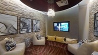 This single-floor space is defined by its distinctive curved architectural design, most notably expressed through a bold, deep burgundy circular ceiling detail that anchors the room's dramatic aesthetic. A cascading crystal chandelier hangs centrally from this curved ceiling element, providing an elegant focal point while casting warm, ambient light throughout the space.

The walls feature a combination of natural stacked-stone textured cladding and smooth taupe-toned surfaces, creating a refined contrast of materials. Two large black-framed abstract artworks are displayed side by side on the stone accent wall, adding a sophisticated artistic element to the interior.

A large wall-mounted flat-screen television is recessed into the smooth wall surface, serving as an entertainment centerpiece. Seating is arranged in a relaxed, lounge-style configuration, including a cream-colored accent chair and a generous sofa adorned with gold and cream cushions, encouraging comfortable viewing and social interaction.

The flooring features a plush carpet with a bold, organic patterned design in warm reds and taupes, reinforcing the space's upscale, lounge-inspired character. Overall, the space conveys a contemporary, luxurious ambiance well-suited for entertaining or relaxation.