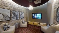 This single-floor space is defined by its distinctive curved architectural design, most notably expressed through a bold, deep burgundy circular ceiling detail that anchors the room's dramatic aesthetic. A cascading crystal chandelier hangs centrally from this curved ceiling element, providing an elegant focal point while casting warm, ambient light throughout the space.

The walls feature a combination of natural stacked-stone textured cladding and smooth taupe-toned surfaces, creating a refined contrast of materials. Two large black-framed abstract artworks are displayed side by side on the stone accent wall, adding a sophisticated artistic element to the interior.

A large wall-mounted flat-screen television is recessed into the smooth wall surface, serving as an entertainment centerpiece. Seating is arranged in a relaxed, lounge-style configuration, including a cream-colored accent chair and a generous sofa adorned with gold and cream cushions, encouraging comfortable viewing and social interaction.

The flooring features a plush carpet with a bold, organic patterned design in warm reds and taupes, reinforcing the space's upscale, lounge-inspired character. Overall, the space conveys a contemporary, luxurious ambiance well-suited for entertaining or relaxation.