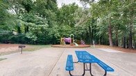 Located at 501 Highway 54 in Carrboro, North Carolina, this outdoor recreational space offers a well-rounded set of amenities set against a lush, wooded backdrop of mature pines and hardwoods. The centerpiece of the space is a vibrant, multi-component playground situated within a circular, wood chip-lined safety border. The play structure features slides, climbing elements, and colorful modular components in purple, green, and orange, designed to engage children of various ages. Adjacent to the playground, a paved patio area provides a convenient gathering spot, complete with a blue picnic table and a nearby grill station, making it well-suited for outdoor dining and social activity. A full-size tennis court enclosed by a black chain-link fence sits alongside the grounds, offering a dedicated space for sport and recreation. The natural landscape plays a significant role in the character of this space — towering pines and deciduous trees frame the perimeter, providing shade and a sense of seclusion. Open grassy areas and pine needle-covered ground connect the various amenity zones, while winding concrete pathways allow easy movement throughout. The wooded edges extend beyond the court and playground, reinforcing the peaceful, nature-integrated atmosphere of this community outdoor space.