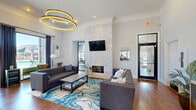 This well-appointed clubhouse in Columbia, SC, offers a thoughtfully designed single-floor space that balances modern aesthetics with a warm, inviting atmosphere. The entryway greets visitors with rich hardwood flooring, soaring ceilings with a distinctive shiplap detail, and a statement large-scale abstract artwork. A round accent table with fresh floral arrangements anchors the welcoming lobby, while a beverage station and reception desk with a bay window provide functional touches.

The living area serves as the centerpiece of the space, featuring a dramatic floor-to-ceiling herringbone-tiled fireplace surround topped by a wall-mounted flat-screen TV. A bold, fluid blue-patterned area rug grounds the seating arrangement of tufted gray sofas around a sleek coffee table. Overhead, an eye-catching double-ring gold pendant light fixture adds a contemporary focal point to the room.

Adjacent to the lounge, a dedicated desk area with large glass sliding doors opens to views of an outdoor pool and surrounding grounds, flooding the interior with natural light. Crown molding throughout, recessed lighting, and a neutral gray-and-white color palette unify the space, creating a polished yet comfortable environment ideal for community gathering and leasing activity.