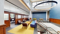 This expansive single-floor workspace masterfully combines historic character with contemporary design sensibilities. The centrepiece is a dramatic double-height atrium flooded with natural light from large clerestory windows, anchored by a striking oversized circular pendant light that commands attention from every angle. Original warm-toned timber panelling and classic wooden partitions with frosted glass panels pay homage to the building's heritage, while blue-painted walls with deliberately preserved raw concrete textures add an industrial, artistic edge.

The lounge area is thoughtfully furnished with mid-century style sofas, armchairs, and wooden coffee tables arranged on vibrant yellow flooring accents set against grey carpet, creating defined zones within the open plan. A dedicated dining or collaborative meeting area features yellow chairs and round tables, offering a colourful contrast to the more neutral tones throughout.

A grand staircase with dark iron balusters and timber handrails connects levels and serves as an architectural focal point. Glass-partitioned private offices or workrooms line one side, maintaining an open and airy feel while providing functional separation. A glazed fitness or activity room is also visible, reinforcing the space's multifunctional nature. Exposed ductwork adds an industrial touch to secondary areas, completing this dynamic, design-forward environment.