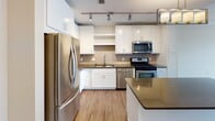 Located on Tenth Ave S in Minneapolis, Minnesota, this contemporary urban residence offers a thoughtfully designed single-floor layout that blends modern finishes with functional living spaces. The open-concept kitchen and living area serves as the heart of the home, anchored by a sleek kitchen island with dark quartz countertops. White shaker cabinetry, a neutral tile backsplash, stainless steel appliances — including a gas range, microwave, and French door refrigerator — and both track and pendant lighting complete the kitchen's polished, practical design. The living area flows seamlessly from the kitchen, featuring durable hardwood-style flooring and large windows that invite abundant natural light and city views. The bedroom is carpeted for comfort and includes a ceiling fan and a generous window overlooking the surrounding neighborhood. A dedicated laundry area houses a stacked washer and dryer unit alongside built-in shelving and a closet, offering excellent storage convenience. The bathroom is clean and bright, finished with light wood-tone tile flooring, a white vanity, and a full tub-shower combination. Neutral tones and contemporary finishes throughout create a cohesive, move-in-ready atmosphere ideal for urban living.