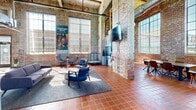 Located at 1700 Fairview St in Greensboro, NC, this single-floor space combines industrial character with modern functionality. The expansive main area is defined by soaring ceilings with exposed wooden beams, visible ductwork, and original brick walls that lend an authentic, raw aesthetic throughout. Terracotta tile flooring runs continuously across the entire floor, adding warmth and cohesion to the industrial design.

Oversized multi-pane windows line the walls, flooding the interior with generous natural light and offering views of the surrounding greenery. The main living area is versatile and well-appointed, featuring multiple seating arrangements, a wall-mounted television, a foosball table, and dining tables with modern chairs — making it ideal for both collaboration and recreation. Informational displays and artwork, including a prominent Josephine Boyd Community Center banner, add cultural character to the space.

A dedicated dining and kitchenette area is equipped with cabinetry, countertop appliances, a sink, and a stainless steel refrigerator, set beneath the same dramatic tall windows. The accessible bathroom retains the brick wall aesthetic while providing ADA-compliant fixtures including grab bars and a comfort-height toilet. Throughout, the space balances its historic architectural bones with contemporary furnishings and practical amenities.