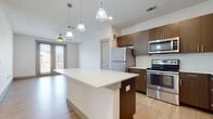This single-floor residence features a thoughtfully designed open-concept layout with a clean, contemporary aesthetic. The kitchen serves as a focal point, outfitted with rich wood-tone cabinetry extending to the ceiling, a white subway tile backsplash, and a full suite of stainless steel appliances including a range, microwave, refrigerator, and dishwasher. A central island with a crisp white countertop provides additional workspace and separates the kitchen from the living area. Pendant lighting hangs above the island, complementing the recessed lighting throughout.

Light hardwood-style flooring runs through the main living and kitchen areas, creating a cohesive and bright atmosphere. The living space is generously sized, with a ceiling fan and access to a glazed exterior door that brings in natural light and outdoor views. A carpeted bedroom offers a quieter retreat, featuring dual windows with neighborhood views and a ceiling fan. The bathroom is finished with full-height white subway tile surrounding the soaking tub, paired with wood-style flooring for a clean, modern look. Overall, the unit presents a well-appointed, contemporary living environment.
