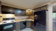 Located at 51 Williamstowne Ct in Buffalo, NY, this single-floor home offers a practical and well-organized layout comprising two bedrooms, one bathroom, a living room, kitchen, and hallway. The kitchen is a standout feature, outfitted with dark wood cabinetry, light countertops, a double-basin sink, and black appliances including a refrigerator, dishwasher, and electric range. Patterned tile flooring runs throughout the kitchen, adding texture to the space. The living room is carpeted and features a sliding glass door dressed with vertical blinds, allowing natural light to filter in and providing access to the outdoors. Both bedrooms are carpeted and finished with warm neutral tones, wood-trimmed doors, and textured ceilings. The primary bedroom includes a window with a view of surrounding greenery, offering a sense of connection to the outdoors. Each bedroom features closet access via interior wood-panel doors. The bathroom is cleanly appointed with a tiled tub-shower combination, chrome fixtures, a toilet, and a vanity with a sink, all set on white tile flooring. A hallway connects the main living areas, completing this efficiently designed home.