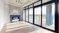 Located in Tulsa, Oklahoma, this thoughtfully designed one-bedroom, one-bathroom unit offers a compelling blend of modern aesthetics and functional living across a single floor. The open-concept kitchen and living room serves as the heart of the space, featuring light hardwood-style flooring throughout, recessed lighting, and a striking fireplace framed by a textured stone tile surround with a navy blue mantle — a true focal point of the room. The kitchen is outfitted with white lower cabinetry, warm wood upper cabinets with glass-front accents, a white subway tile backsplash, and light stone countertops, delivering a clean, contemporary look. Floor-to-ceiling black-framed glass doors flood the living area with natural light and open directly onto a private balcony, which offers expansive open-air views of the surrounding Tulsa landscape. The bedroom is a calm retreat with light hardwood flooring, neutral grey walls, a ceiling fan, and a dedicated closet. The bathroom is a standout space, showcasing polished marble-look tile floors, a full-height marble-tiled walk-in shower with gold-accented frameless glass sliding doors, and a sleek floating vanity — all finished with warm wood door accents and gold hardware throughout.