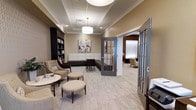 Located at 659 Rice Boulevard in Exton, PA, this single-floor professional space is designed with a polished, welcoming aesthetic that balances comfort and sophistication. The reception and waiting area serves as the centerpiece of the space, featuring warm neutral tones, a tray ceiling with crown molding, and a combination of recessed lighting and a statement pendant fixture that create a bright, inviting atmosphere. Seating options include upholstered armchairs, ottoman stools, and a cushioned bench-style sofa, all arranged to foster a relaxed and professional environment. A built-in dark-wood bookcase lines one wall, adding both function and a refined design element. Tasteful artwork adorns the walls, complementing the neutral color palette. French-style glass-paned interior doors partition the waiting area from adjacent rooms, allowing light to flow throughout while maintaining a sense of privacy. Glimpses into adjoining spaces reveal additional seating areas, suggesting a layout well-suited for client-facing professional services. The overall ambiance is calm, clean, and carefully curated, projecting a professional yet approachable atmosphere throughout.