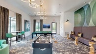 Located at 4535 SW 54th St Rd in Ocala, Florida, this contemporary game room is a well-appointed recreational space designed for social engagement and entertainment. The room is anchored by a bold, abstract carpet in muted blue and grey tones that grounds the energetic atmosphere. A full-size pool table and a foosball table serve as the centerpieces, complemented by a round poker or game table surrounded by mint-green upholstered chairs. A built-in banquette finished in soft lavender-grey provides comfortable lounge seating, accented by gold-toned throw pillows and sculptural wooden side tables. The walls feature striking artistic touches, including a large-scale geometric mural in sage green and gold, a vibrant horse portrait, and a green moss wall visible through a connected lounge area beyond. Tall windows dressed with flowing textured drapes allow ample natural light and offer views of an outdoor pool area. A wall-mounted flat-screen TV adds a media component to the space. Overhead, a dramatic sculptural chandelier composed of interlocking brass and frosted rings serves as a standout design statement. The overall ambiance is modern, curated, and inviting, blending recreational function with upscale interior design.