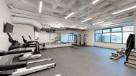 This fitness center offers a spacious, open-plan layout designed to accommodate a wide range of workout activities. The space is finished with durable speckled rubber flooring throughout, providing a safe and practical surface for exercise. Elevated ceilings with an exposed structural grid are illuminated by rows of linear fluorescent lighting, ensuring bright and even coverage across the entire floor.

The main workout area is divided into functional zones. A cardio section features multiple treadmills, each paired with a wall-mounted flat-screen television for entertainment during workouts. Adjacent to this area, a dumbbell rack offers a comprehensive selection of free weights, complemented by kettlebells and additional accessories. A multi-station cable machine and weight bench provide dedicated strength training options, while a Pilates reformer adds versatility for low-impact exercise.

Full-length mirrors line one wall, enhancing the sense of depth and allowing users to monitor their form. Large windows along an exterior wall invite natural light into the space and offer views to the outside. A staircase with black metal railings leads to an exit, ensuring convenient access. The overall aesthetic is clean and functional, with white walls and an industrial-style ceiling that give the space a modern, professional character suited to a variety of fitness needs.