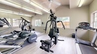 Located at 7566 Greenback Ln in Citrus Heights, CA, this dedicated exercise room offers a well-equipped fitness space designed for convenient, on-site workouts. The room features light wood-style flooring and soft, neutral-toned walls accented by a vibrant teal blue feature wall, creating an energizing yet welcoming atmosphere. A ceiling fan overhead provides air circulation, complemented by fluorescent lighting that keeps the space bright and functional. Large, multi-pane windows line one wall, flooding the room with natural light and offering pleasant views of an outdoor pool area and surrounding greenery — an inviting backdrop for any workout. A mirrored wall runs along one side of the room, visually expanding the space and allowing users to monitor their form during exercise. The fitness equipment includes a multi-function cable strength training machine, a treadmill, and an elliptical trainer, providing options for both cardiovascular and resistance training. A paneled entry door with a glass insert offers easy access to the exterior. Informational postings are displayed on the wall for user reference. The overall design is clean, practical, and well-maintained, making this exercise room a functional amenity for residents and users of the Citrus Heights community.