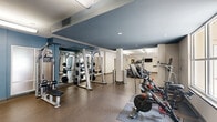 Located at 2101 Arlington Blvd in Charlottesville, Virginia, this well-appointed fitness center offers a generous, open floor plan designed to accommodate a wide variety of workout routines. The space is thoughtfully divided into distinct exercise zones, each equipped to support different training needs.

The cardio area features a row of modern treadmills, elliptical trainers, and stationary cycling bikes, all positioned to take advantage of large windows that allow ample natural light and provide views of the surrounding outdoor areas. The bright, airy atmosphere is further enhanced by overhead fluorescent lighting that ensures consistent illumination throughout.

A dedicated free weights zone includes a full rack of dumbbells in graduated sizes, adjustable weight benches, and ample open floor space for stretching and floor exercises. This section is complemented by a wall-mounted television for entertainment during workouts.

The functional training area is stocked with medicine balls, kettlebells, resistance equipment, and a cable machine, offering versatility for dynamic and strength-based exercises. Durable, cushioned flooring throughout provides comfort and safety underfoot.

The interior design features a clean, modern aesthetic with neutral tones accented by slate-blue walls and white ceilings. Structural columns define the space without interrupting the open feel. Lush decorative plants near the entrance add a welcoming touch to this comprehensive, well-equipped exercise facility.