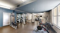 Located at 2101 Arlington Blvd in Charlottesville, Virginia, this well-appointed fitness center offers a generous, open floor plan designed to accommodate a wide variety of workout routines. The space is thoughtfully divided into distinct exercise zones, each equipped to support different training needs.

The cardio area features a row of modern treadmills, elliptical trainers, and stationary cycling bikes, all positioned to take advantage of large windows that allow ample natural light and provide views of the surrounding outdoor areas. The bright, airy atmosphere is further enhanced by overhead fluorescent lighting that ensures consistent illumination throughout.

A dedicated free weights zone includes a full rack of dumbbells in graduated sizes, adjustable weight benches, and ample open floor space for stretching and floor exercises. This section is complemented by a wall-mounted television for entertainment during workouts.

The functional training area is stocked with medicine balls, kettlebells, resistance equipment, and a cable machine, offering versatility for dynamic and strength-based exercises. Durable, cushioned flooring throughout provides comfort and safety underfoot.

The interior design features a clean, modern aesthetic with neutral tones accented by slate-blue walls and white ceilings. Structural columns define the space without interrupting the open feel. Lush decorative plants near the entrance add a welcoming touch to this comprehensive, well-equipped exercise facility.