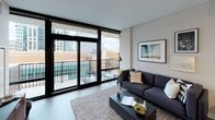 This thoughtfully designed single-floor residence offers a cohesive modern aesthetic with clean lines and a neutral color palette throughout. The open-concept living and kitchen area forms the heart of the unit, where the kitchen stands out with white flat-front cabinetry extending to the ceiling, dark quartz countertops, white subway tile backsplash, stainless steel appliances including a gas range and French door refrigerator, and a peninsula with bar seating. Track lighting overhead enhances the functional workspace. The living area flows naturally from the kitchen, providing a comfortable setting with direct access to a private balcony through floor-to-ceiling sliding glass doors framed in matte black. The balcony offers sweeping urban skyline views. The bedroom continues the floor-to-ceiling window theme, flooding the space with natural light and showcasing elevated city views. Soft carpet underfoot contrasts the hardwood-style flooring found in the main living areas. The full bathroom features a white vanity with ample storage, a tub-shower combination clad in white subway tile, and bright, clean finishes. A dedicated in-unit laundry closet with a stacked washer and dryer adds everyday convenience. A secondary half bath provides additional functionality.