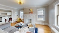 This single-floor residence showcases a thoughtful blend of traditional character and contemporary updates throughout its well-proportioned layout. The living room serves as a focal point, anchored by a white-painted brick fireplace flanked by double-hung windows that flood the space with natural light. Crown molding and hardwood floors carry through the common areas, lending a cohesive, classic aesthetic. A striking sputnik-style chandelier adds a mid-century modern accent overhead.

The dining area accommodates a glass-top table and sits beside a pair of windows with a cast-iron radiator beneath, reinforcing the home's period charm. Adjacent, the kitchen is outfitted with white shaker-style cabinetry, marble-look countertops, a subway tile backsplash, and stainless steel appliances including a gas range with wall-mounted hood, refrigerator, and dishwasher.

The bedroom features hardwood flooring, crown molding, and direct access to a bathroom with classic black-and-white mosaic tile flooring and in-unit laundry. A dedicated office space with a large window provides a bright, functional work area. Neutral tones throughout create a calm, cohesive ambiance suited to comfortable everyday living.
