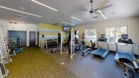 Located at 6811 E Main St in Scottsdale, Arizona, this single-floor fitness center is thoughtfully designed to support a comprehensive workout experience. The space features a vibrant interior with a distinctive color palette of yellow, teal, and white horizontal stripes along the accent wall, lending an energetic and modern aesthetic throughout.

The cardio zone is well-appointed with treadmills, elliptical machines, a stair climber, and a stationary bike, all positioned near large windows that allow abundant natural light and views of the surrounding outdoor area. Ceiling fans provide additional airflow and comfort during workouts.

The strength training section offers a multi-functional cable machine, a dedicated weight bench, and a fully stocked dumbbell rack ranging across multiple weight increments. Floor-to-ceiling mirrors along one wall enhance the sense of space and allow for proper form checks during exercise.

A separate functional training area houses resistance equipment, barbells, and stability balls, catering to a variety of fitness routines. Built-in cabinetry and a water fountain provide convenient storage and hydration options. Wall-mounted televisions add an entertainment element to cardio sessions.

Double glass doors open directly to an outdoor courtyard area, seamlessly connecting the fitness center to the exterior amenities of the property.