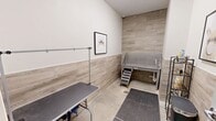 This state-of-the-art dog spa in Marlborough, MA, spans 210 square feet on a single floor, offering a tailored environment for canine grooming and care. The facility boasts two distinct areas, each designed for specific pet services. A long, well-lit corridor with polished concrete flooring leads to various treatment rooms, creating an organized and efficient layout. The walls are painted in neutral tones, primarily grey, lending a calm and professional atmosphere. One room is equipped with a large, elevated stainless steel grooming table accessible via steps, ideal for bathing and grooming services. This space features wood-look tiled wainscoting, adding warmth to the clinical setting. Thoughtful touches include framed dog artwork, a multi-tiered supply cart, and strategically placed grooming tools. The spa's design emphasizes cleanliness and functionality, with ample lighting and easy-to-clean surfaces throughout, ensuring a hygienic environment for pets and groomers alike.