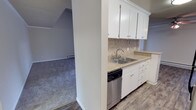 This single-floor space is thoughtfully designed with a practical and clean aesthetic. The kitchen is a standout feature, showcasing white shaker-style cabinetry that extends to the upper walls, providing generous storage. A neutral stone-toned countertop complements the classic subway-style tile backsplash, while a stainless steel double-basin sink and a stainless steel dishwasher add functional, modern touches. Wood-look flooring runs through the kitchen area, offering durability and a warm visual contrast to the bright cabinetry.

A half-wall separates the kitchen from the adjacent living area, creating a sense of openness while maintaining a defined layout. The living area features plush carpeting in a neutral grey tone, contributing to a comfortable and cozy atmosphere. Vaulted ceilings in the living space lend an airy, expansive feel, and a baseboard heater along the far wall ensures year-round comfort. Crown molding details add a subtle architectural refinement throughout. The overall design strikes a balance between practicality and comfort, making it a functional and welcoming space.