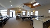 This single-floor community space is designed to accommodate a variety of social and recreational activities, combining thoughtful architectural details with a warm, welcoming ambiance. The centerpiece of the main room is a full-size pool table positioned beneath a striking vaulted ceiling with a coffered accent panel in a rich warm tone, adding visual depth and character to the space. Recessed lighting and a ceiling fan ensure the area is both well-lit and comfortable year-round. A gas fireplace flanked by wall sconces anchors a cozy lounge area furnished with plush seating, creating an inviting spot for relaxed conversation. Large windows and glass double doors flood the interior with natural light while offering views to the outdoors. A bar-height counter with granite countertops and bar stool seating lines one side of the room, ideal for casual gatherings. A dedicated dining area with high-top tables and chairs provides additional seating. Elegant finishing touches, including a chandelier and decorative artwork, lend a refined character throughout. The open layout promotes a seamless flow between each zone, making this a versatile and well-appointed gathering space.