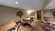 This single-floor community space is designed to foster a comfortable and social atmosphere. The main gathering area is generously sized, featuring patterned carpeting throughout and a neutral warm color palette on the walls. Multiple round tables with cushioned chairs are thoughtfully arranged to encourage group interaction and flexible use of the space. Decorative touches, including a striking metal tree wall sculpture and ornamental wall pieces, lend character and warmth to the room.

Natural light filters in through a window fitted with vertical blinds, complemented by recessed ceiling lighting and a floor lamp that together create a welcoming ambiance. A wall-mounted television adds a functional element for shared viewing.

An interior pass-through window connects the main room to a compact kitchenette, which is equipped with a range, white cabinetry providing ample storage, and countertop workspace. This kitchen area is well-suited for preparing and serving refreshments during gatherings. A decorative wall clock adds a charming accent near the pass-through.

The space also features a corridor leading to additional rooms, enhancing its versatility. Overall, this thoughtfully appointed community room offers a practical and inviting setting for social events, meetings, and everyday use.