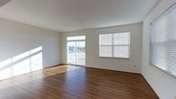 This three-floor space offers a clean, modern aesthetic defined by crisp white walls and rich, dark hardwood-style flooring that runs throughout. The main living area is generously proportioned, featuring an open layout that allows natural light to pour in from multiple sources. A sliding glass door leads directly to a private balcony, offering outdoor views and additional brightness that enhances the airy feel of the room. A adjacent window fitted with horizontal blinds provides further light control and ventilation options.

The flooring makes a strong visual statement, with warm, dark tones that contrast effectively against the neutral white walls and ceiling, creating a grounded yet bright atmosphere. The ceiling height adds to the sense of openness, contributing to the overall spacious feel of the living area.

The space maintains a neutral, versatile palette throughout, making it well-suited for a variety of interior styles. Spread across three floors, the layout provides ample opportunity for dedicated living, sleeping, and functional zones. The combination of natural light, outdoor access, and clean architectural lines gives this space a welcoming and practical character.